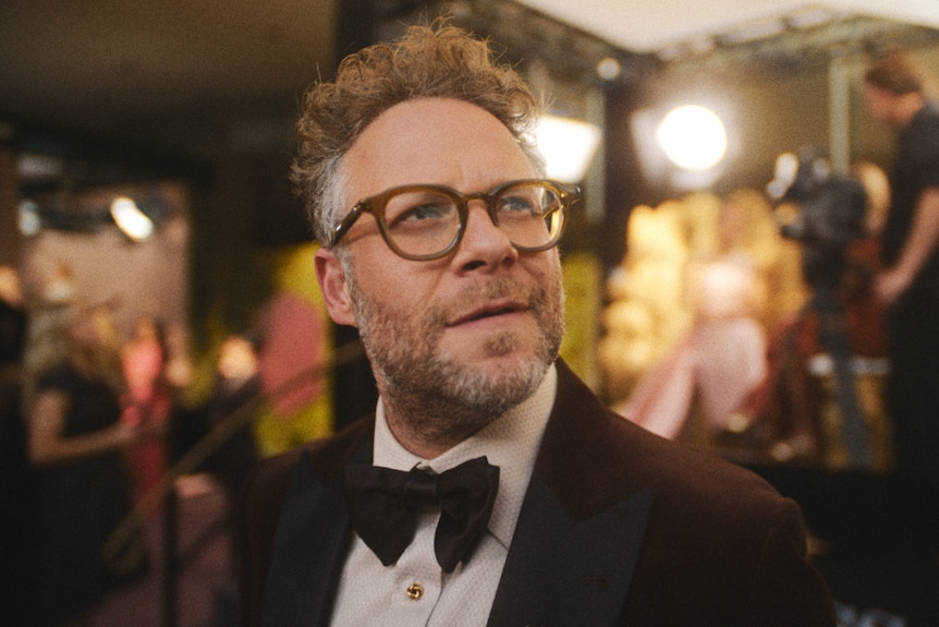 A TV still of Seth Rogen, 43, wearing glasses, head tilted upward in thought, on a red carpet, a person filming behind him.