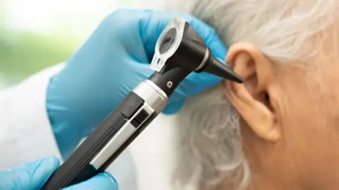 Getty Images A close-up of a medical examination where a healthcare professional is inspecting a person's ear using an otoscope. The healthcare professional is wearing blue disposable gloves. The person being examined has short, grey hair. The background is softly blurred, keeping the focus on the ear and the otoscope.