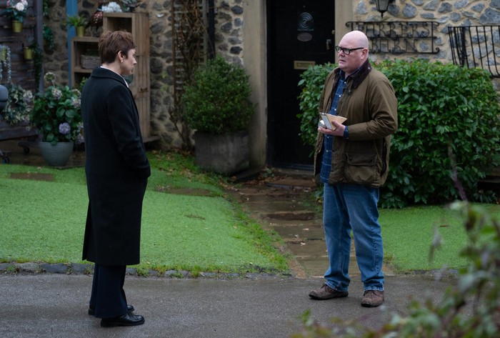 Paddy, holding a sandwich, stands talking to DS Walsh in the village in a scene from Emmerdale