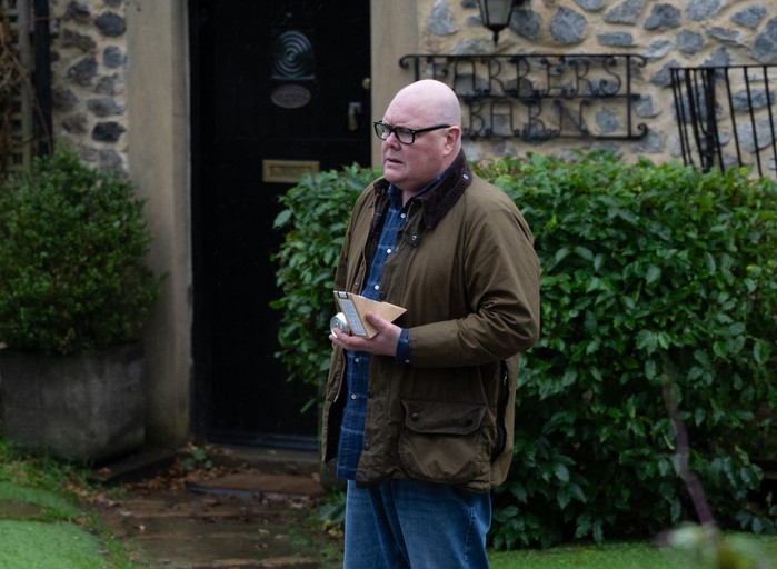 Paddy stands alone, holding a sandwich, in a scene from Emmerdale village