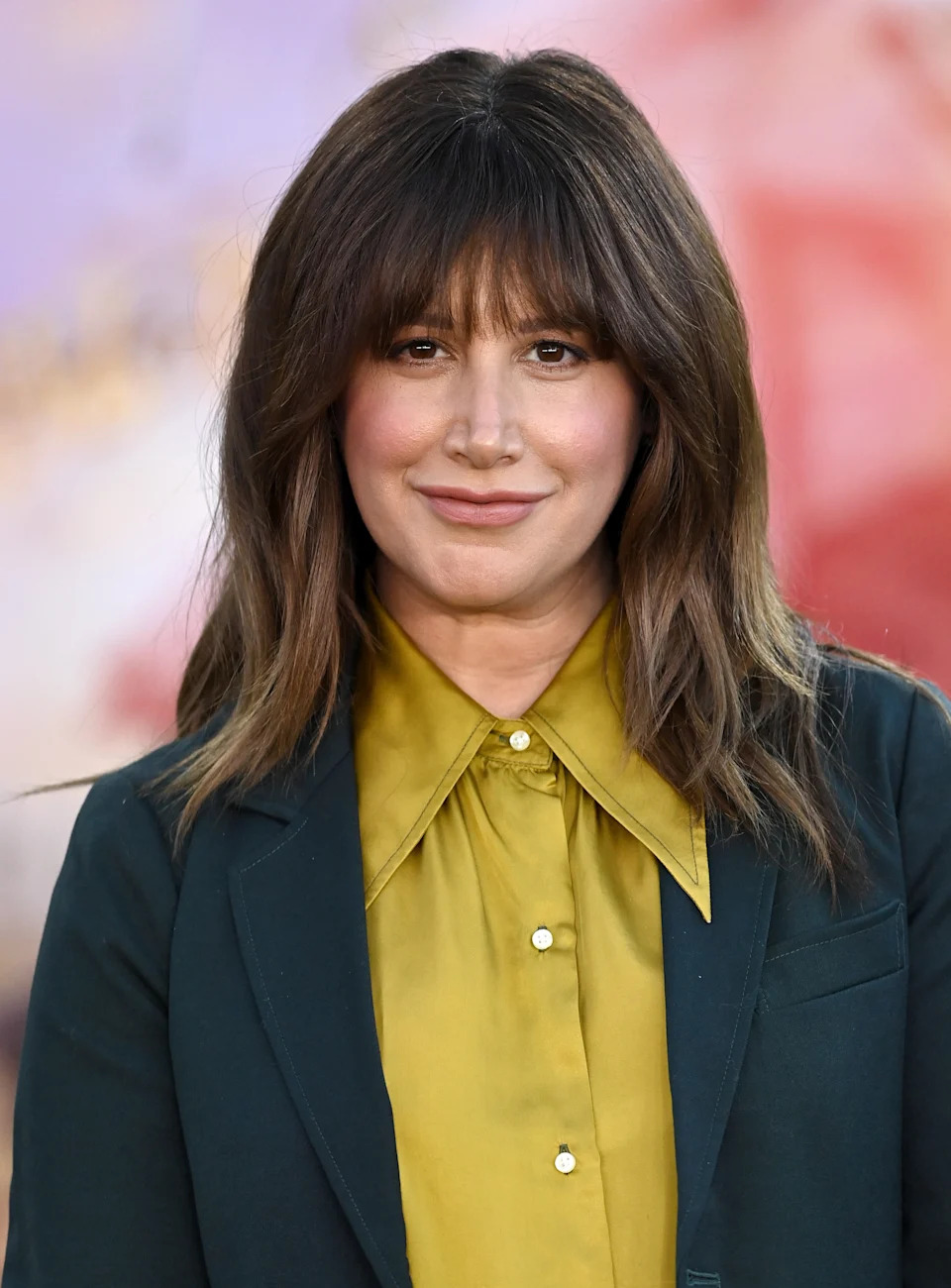 A woman with long hair and bangs smiles on the red carpet, wearing a tailored dark jacket over a buttoned shirt