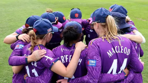 Essex County Cricket Club The Essex women's side link arms to huddle together on the field. They are wearing purple kits and navy caps.