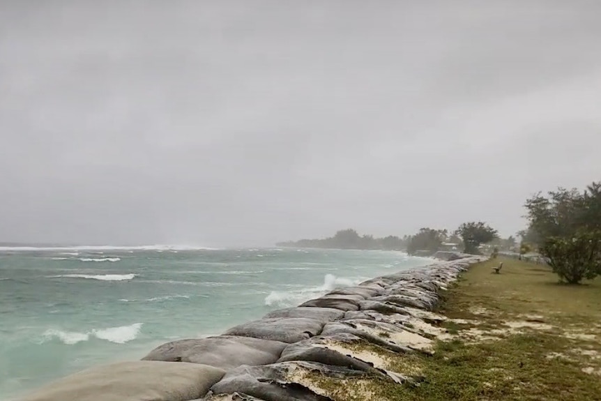 grey skies with blue waters that are rough, sandbags are placed along the water