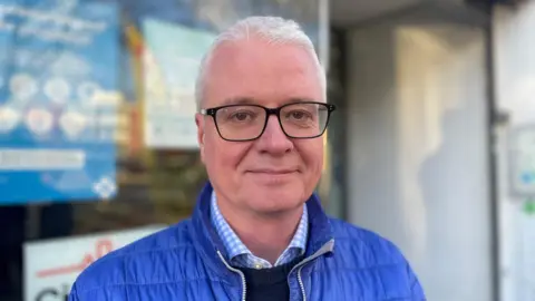 A man with close cropped white hair and black glasses. He is wearing a blue puffer jacket. 