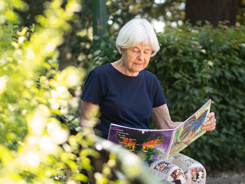 An older woman with short, white hair reads a book while she sits in a park.