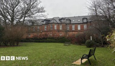 A red brick building with many windows behind a small park with benches, trees and hedges.