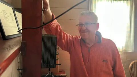 National Trust Geoff has short grey hair and is wearing a red jumper. He has his hand resting on some of the radio equipement.