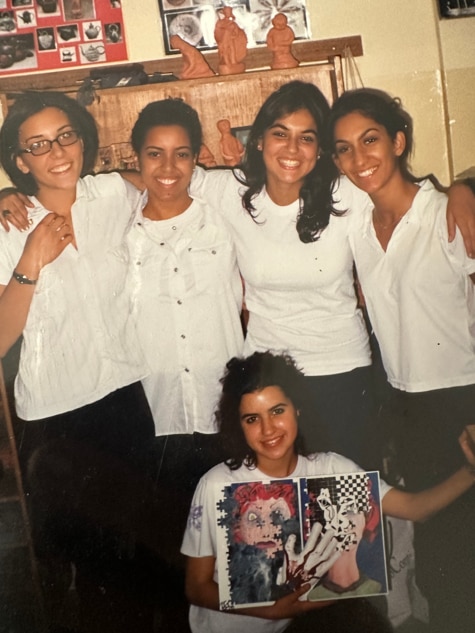 Yasmine, bottom centre, displays a portfolio with art in it while in a uniform crouched below a group of four other students.