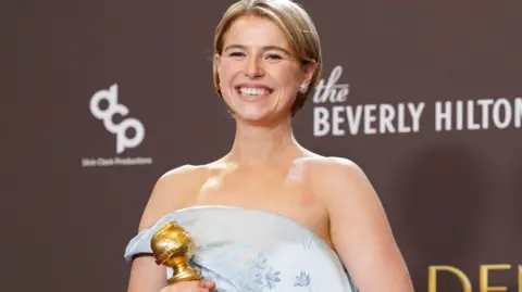 REUTERS/Mario Anzuoni Jessie Buckley, a woman with short, blonde hair, poses with her Golden Globe award.  She is wearing an off the shoulder silver/pale blue gown. 