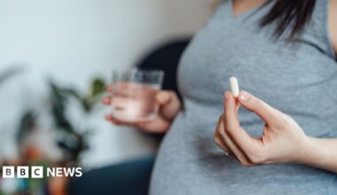 A pregnant women wearing a grey top is visible from the chest down, and holds a white pill in her left hand and a glass of water in her right (slightly blurred)