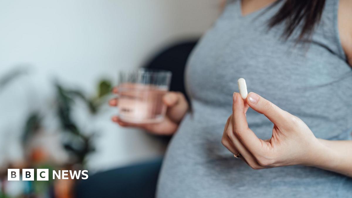 A pregnant women wearing a grey top is visible from the chest down, and holds a white pill in her left hand and a glass of water in her right (slightly blurred)