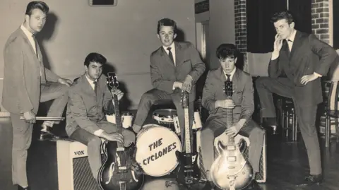 Ricky Ford Five young men photograhped in the 1950s wearing suits typical of that period. Two are holding guitars, one is standing over a drumkit with "The Cyclones" written on the front.