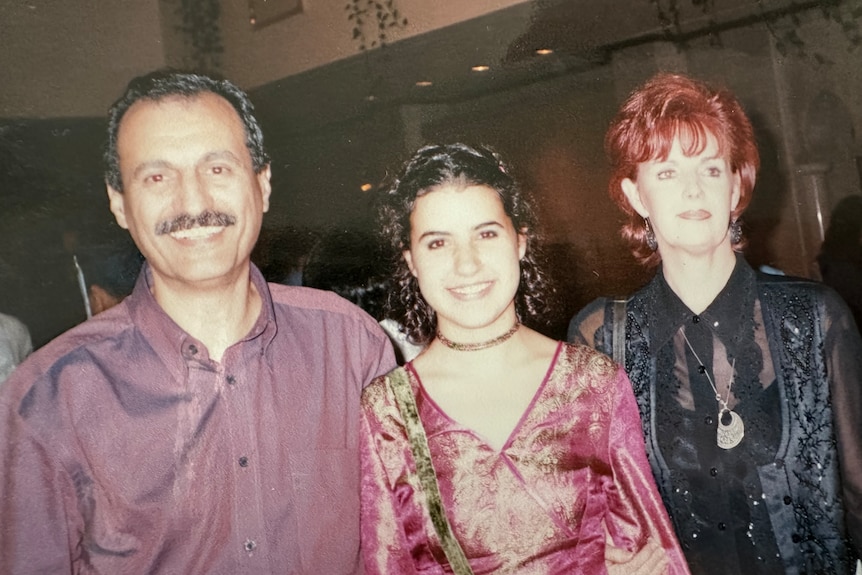 Yasmine smiles with her dad on her right and her mum on her left in a washed out retro photo taken at a formal event.
