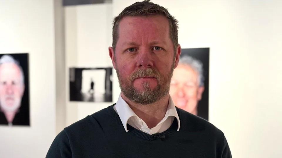 Neil Irwin is standing in a gallery - there are pictures of people in the background. He is wearing a white shirt and dark jumper. He has a beard and brown hair.