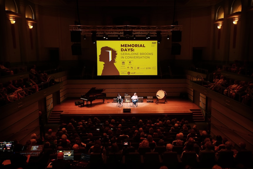 In a concert hall, Geraldine Brooks, 70, and Rosemarie Milsom, middle-aged, sit on stage, in front of a grand piano. 