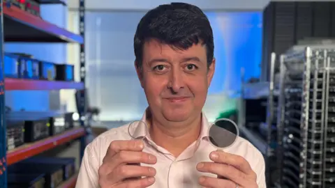 Dr Paul Cain in an engineering lab with racks of equipment on either side lit by blue light. He is wearing a pale pink shirt and holding a round lens up to the camera in each hand. One is transparent, the other is tinted dark grey.