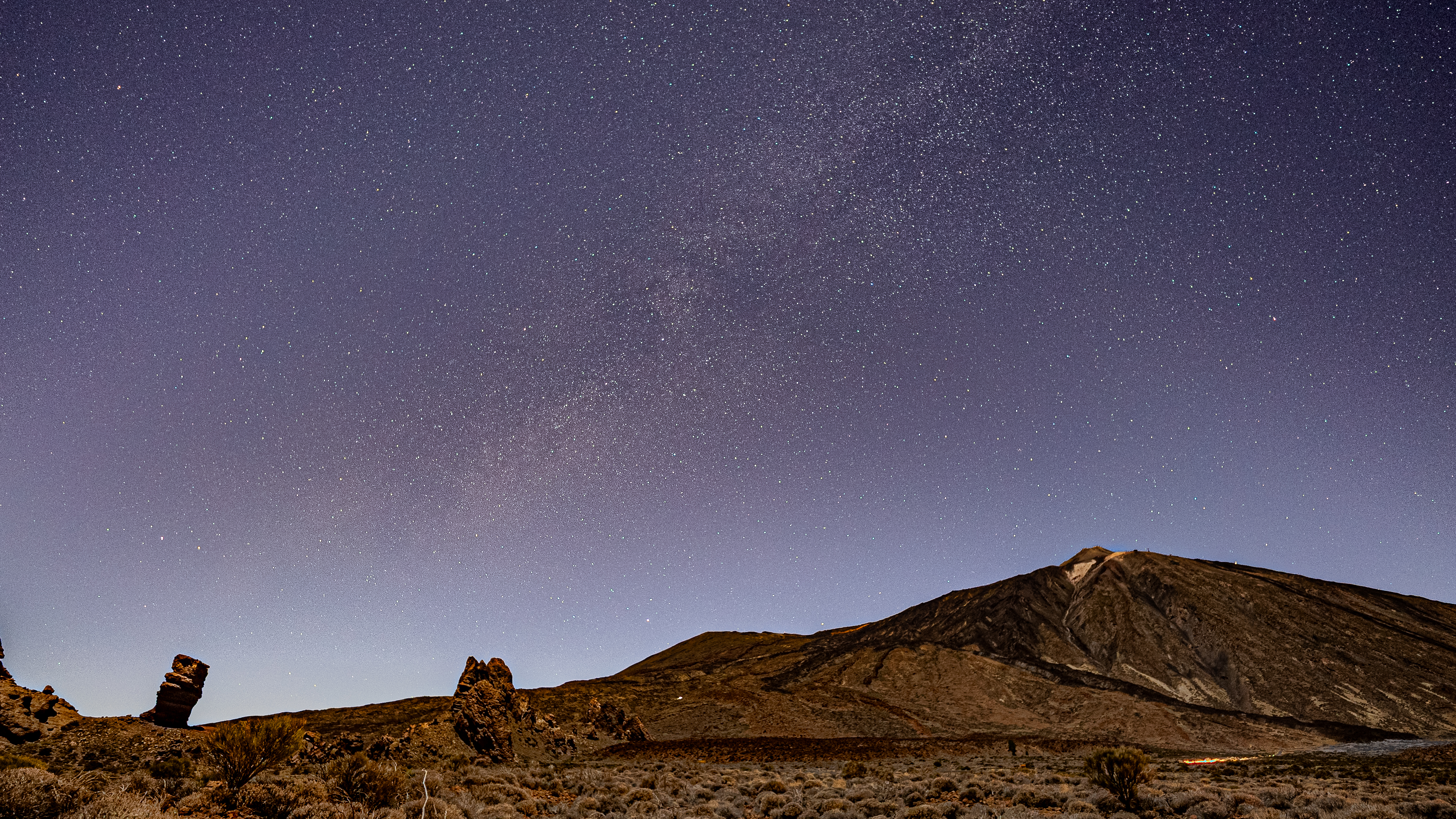 rock formations in the foreground, a starry night sky in the background and a distant volcano.