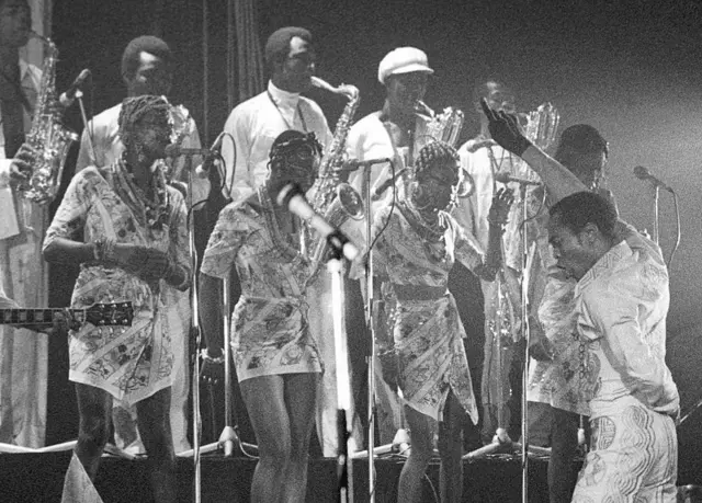 Black and white foto of Fela Kuti wia e dey back di audience and face im back-up singers and band. E dey sing wit one hand up dey point finger while di oda hand dey hide for im back wia e pose as torero.