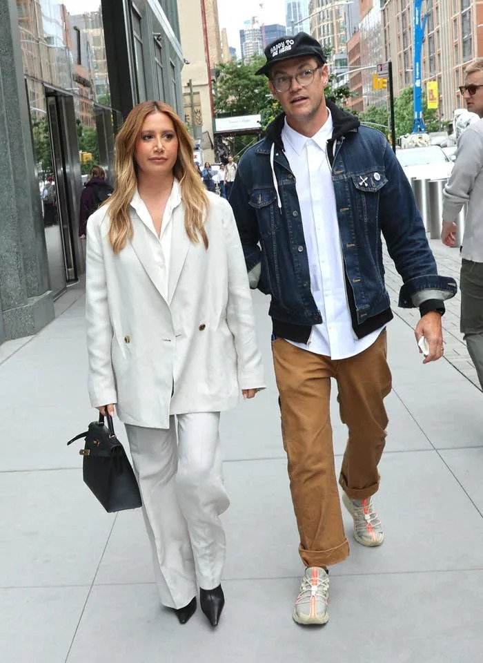Two people walking on a city street; one in a white suit with a black handbag, the other in a denim jacket and cap, wearing casual brown pants