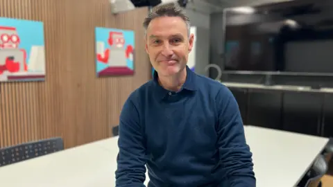 Warren Mauger pictured with short grey and brown hair. He is wearing a navy long-sleeved sweater with a navy shirt collar poking out the top.