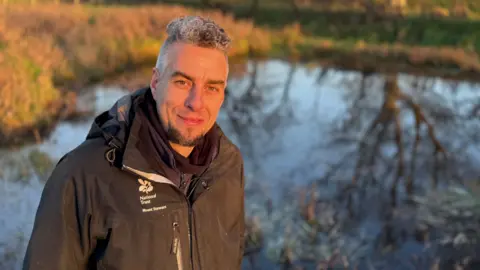 BBC Toby Edwards has short grey and black hair and a black beard. He is standing in front of a pond and is wearing a black coat.