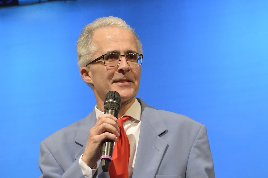 Neil Wright wears a suit jacket and glasses and speaks into a microphone.
