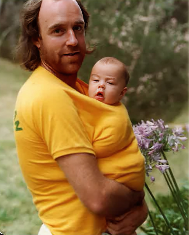 A man holds a baby inside his shirt while standing in a garden.