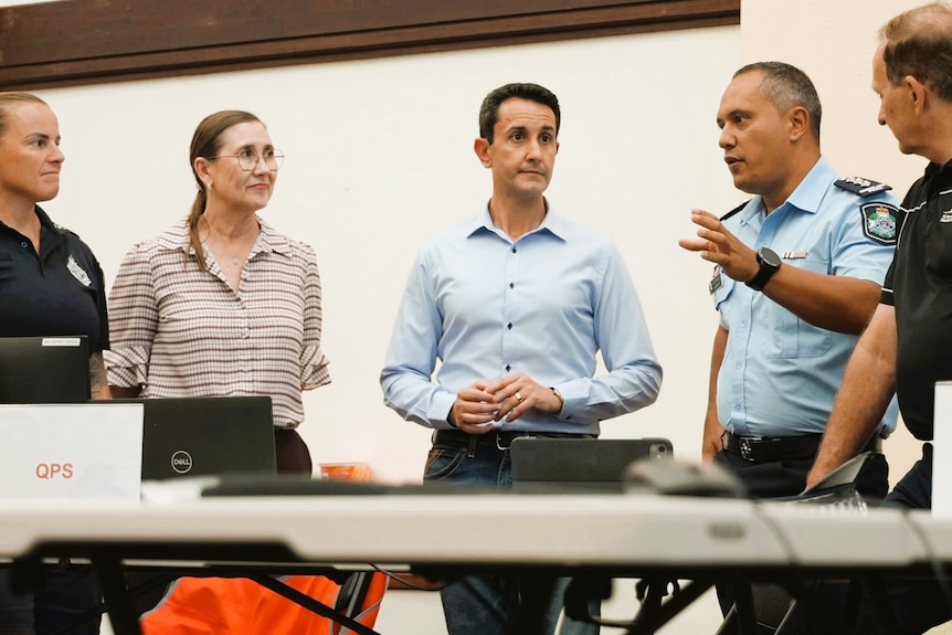 A man in a button up shirt indoors surrounding by four other people listening intently 
