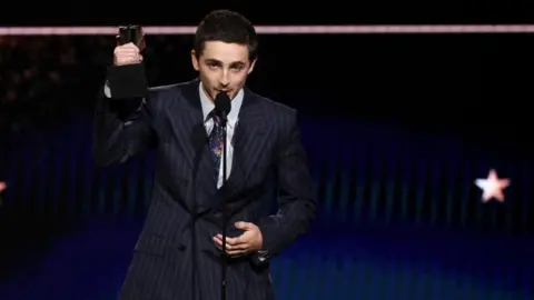 Reuters Timothée Chalamet, who is wearing a pin-striped suit, white shirt and patterned tie, accepts the Best Actor Award for Marty Supreme at the 31st Annual Critics Choice Awards in Santa Monica, California. He is holding his award in the air as he talks into a microphone. 