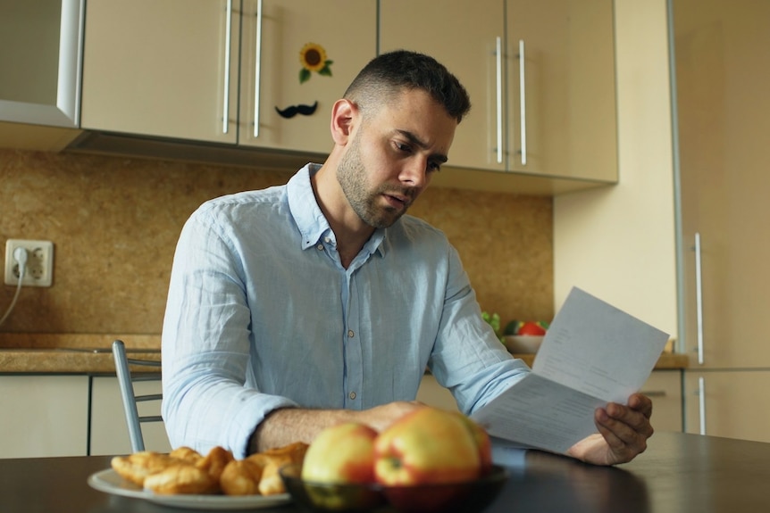 A concerned looking man sits at a table in a kitchen looking at bill.