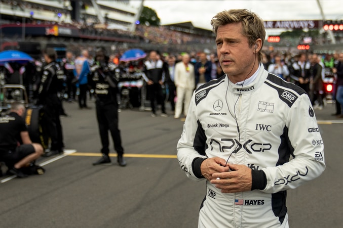 Brad wears a white and black racing suit, headphones in, as he stands on the track with tens of crew and bystanders behind him.