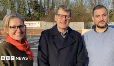 A woman with blonde hair wearing glasses and a green jumper and red scarf smiles towards the camera, she is standing next to a man with grey hair smiling toward the camera. He is wearing glasses and a dark jacket and is standing next to a a bearded man with dark hair who is smiling toward the camera. He is wearing a grey knitted jumper. All three are stood in a line in a car park , wooden fencing and trees can be seen in the background.