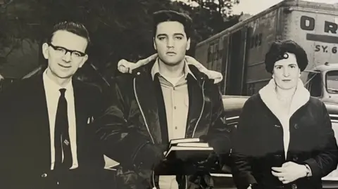 A black and white photograph of three people. The man in the centre is Elvis Presley wearing a shirt, a black, fleeced leather jacket and black gloves. He is holding three books. The man to the left has black hair, black rimmed glasses, a white shirt and a black tie. The woman on the right has dark short hair and a dark fleeced coat. Behind them is a van, a car and trees.