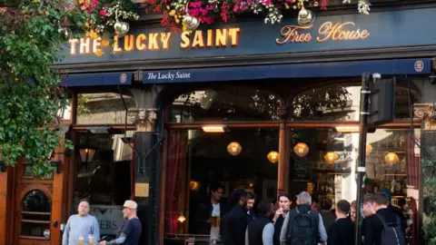 Lucky Saint A pub with punters outside covered in flowers and woodwork. Many people stand outside with pints.