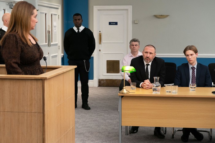 Vicki, stood in a witness box, staring downwards at Ross and Joel, who are sitting in a court room in a scene from EastEnders
