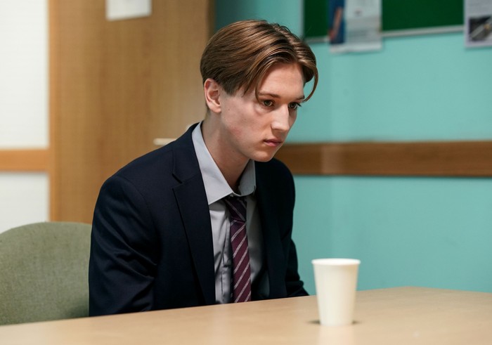 545918,TITLE:Eastenders Joel, wearing a blue suit and burgundy tie, looks upset in front of a white cup on a table in a scene from EastEnders