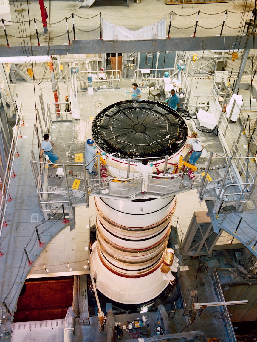 Inside a large construction facility, a white rocket is half built with engineers working on its top surface from scaffolding.