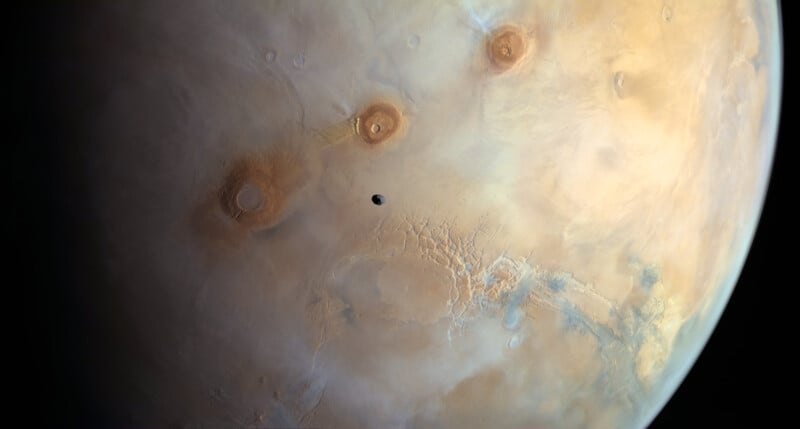 A detailed image of Mars shows its reddish, cratered surface with three large volcanoes near the center and a small, dark moon casting a shadow as it passes overhead.