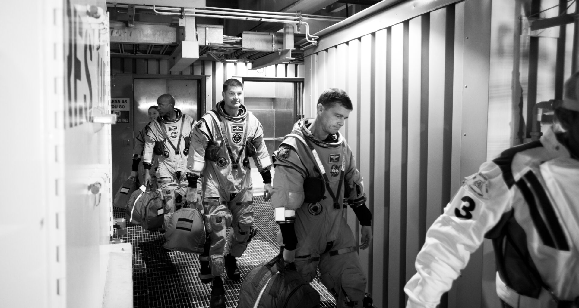 Four astronauts in spacesuits walk through a small corridor, following another person. The photo is in black and white.
