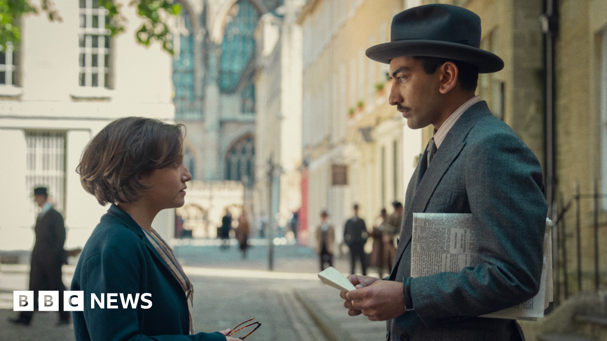 A young woman stands in profile on the left, wearing a blue coat, looking up at a man wearing a grey suit and hat, with a newspaper under his arm. They are stood on a regency-era-style street.