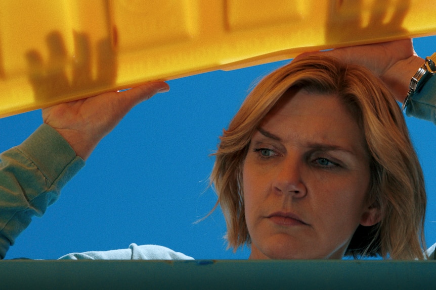 A blond woman peaks into a dumpster with a yellow lid