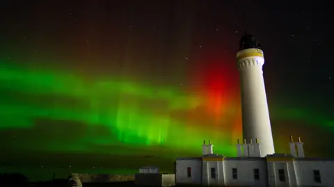 Scott Mellis A tall white lighthouse stands beside low buildings under a vivid night sky lit by green and red aurora. Stars are faintly visible above the glowing lights.