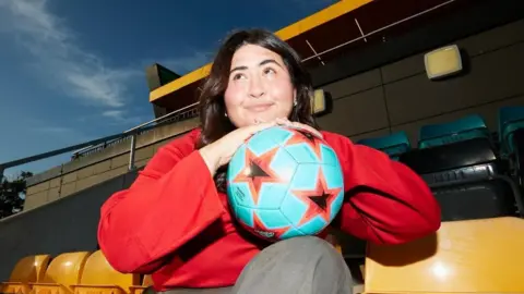 Dani Gonçalves Dani is hold a pale blue football with orange stars. She has shoulder length black hair and brown eyes. She is wearing a red long sleeved top sat on a yellow football stadium seat. Behind her you can see the other empty seats and a bit of blue sky in the left top corner. 
