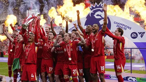 PA Wire Liverpool Football Club celebrate winning the Premier League title. A group of players in red wearing medals jump in the air, lifting a silver trophy. In the background we can see the stadium, with fireworks and a banner that reads 'Champions 2024/25'.
