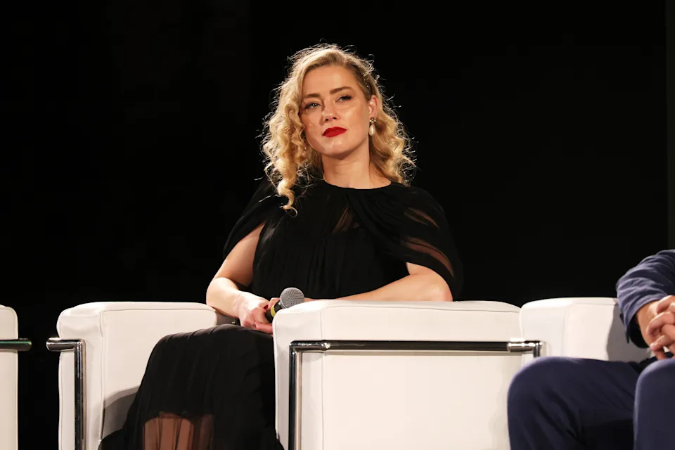 Amber Heard with wavy hair in an elegant black dress sits on a white chair while holding a microphone during a panel discussion