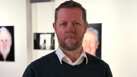 Neil Irwin is standing in a gallery - there are pictures of people in the background. He is wearing a white shirt and dark jumper. He has a beard and brown hair.