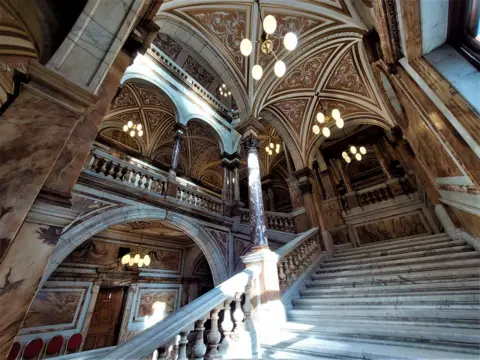 Victor Tregubov An ornate marble staircase rises toward arched, intricately decorated ceilings. Hanging lights illuminate the grand interior, highlighting carved pillars and detailed architectural patterns.
