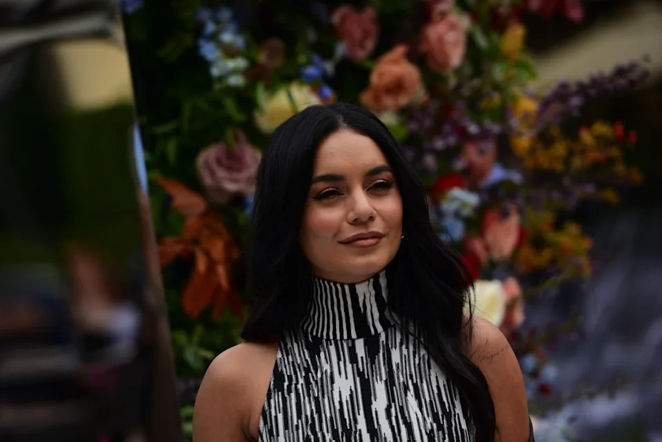 Vanessa Hudgens with long hair and a high-neck patterned dress poses in front of a floral backdrop