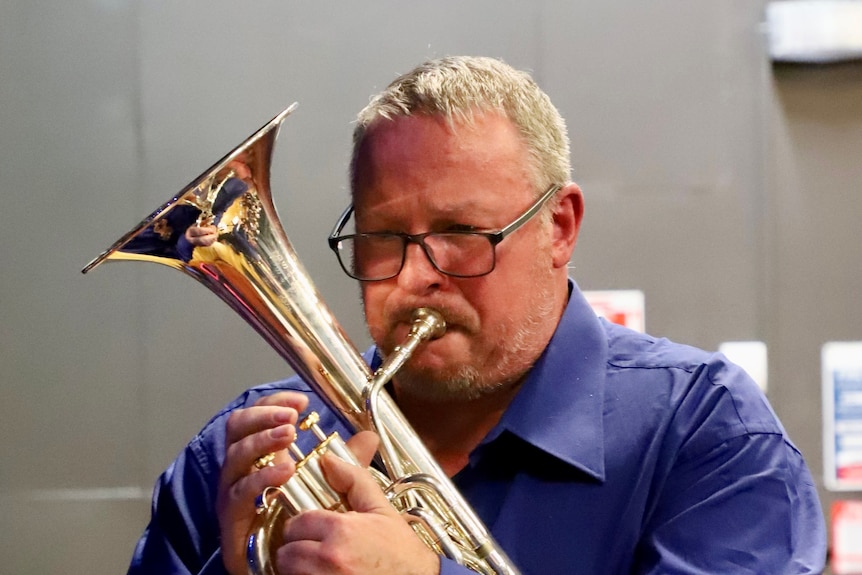Jeff Markham has glasses and short hair, and plays a brass instrument.