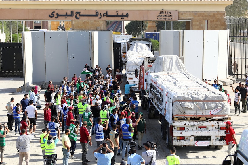 A crowd of people, some wearing hi-vis vests, gather beside several trucks loaded with covered goods as they emerge from a wall.